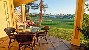 A covered patio with wicker chairs and a glass table, overlooking a golf course and buildings.
