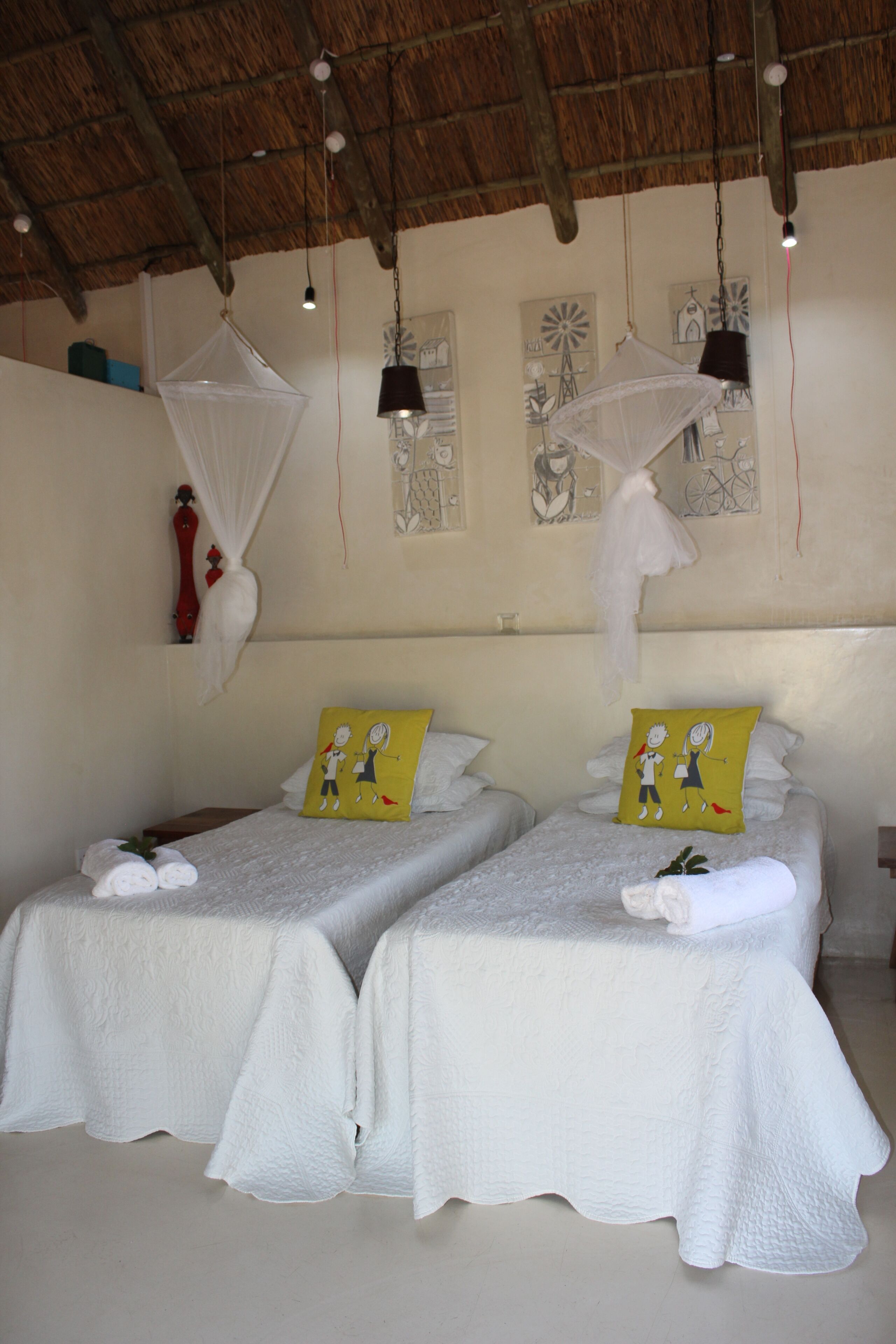 Una habitación con dos camas, cada una con una colcha blanca y un cojín amarillo. Hay luces colgantes y una mosquitera sobre las camas.