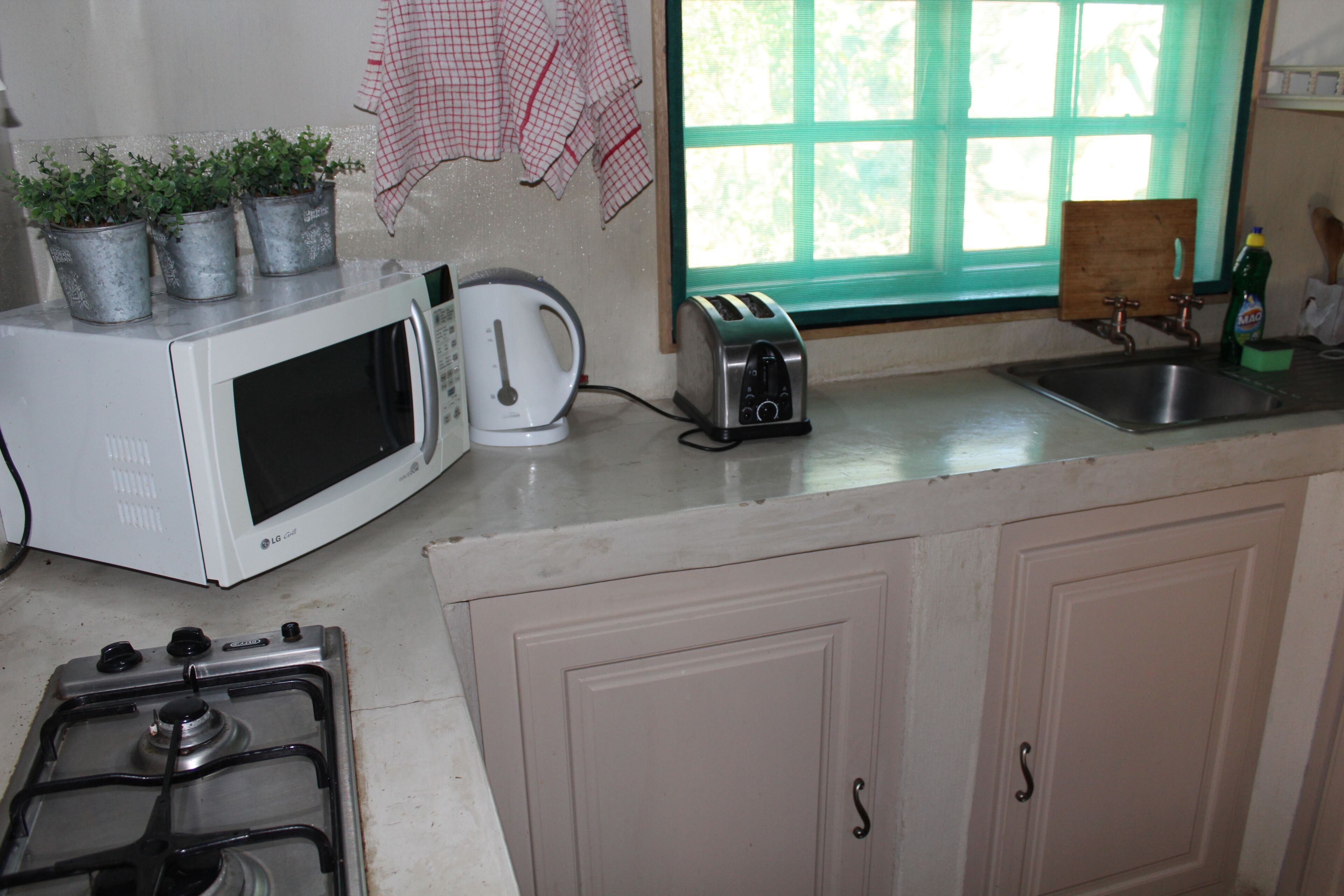 A kitchen with a microwave, kettle, toaster, and stove. There are potted plants on the microwave, a window with a view, and a wooden cutting board on the counter.