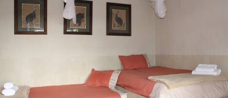 Habitación de hotel con dos camas, cada una con cubrecama rojo y beige, y una mesita con toallas. En la pared hay tres cuadros enmarcados de un pájaro.
