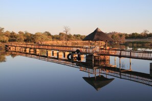 A wooden bridge over a calm body of water, a thatched-roof structure, and a reflection of the surroundings.