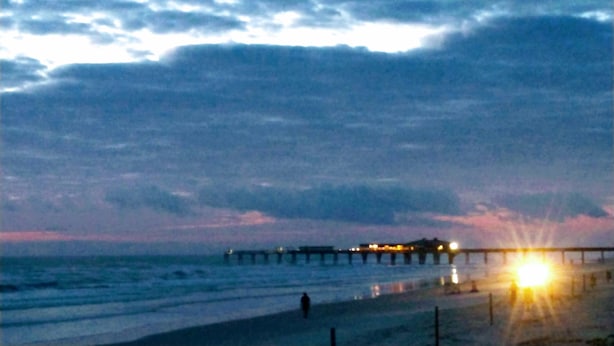 Un muelle que se adentra en el mar, con una persona caminando por la playa.