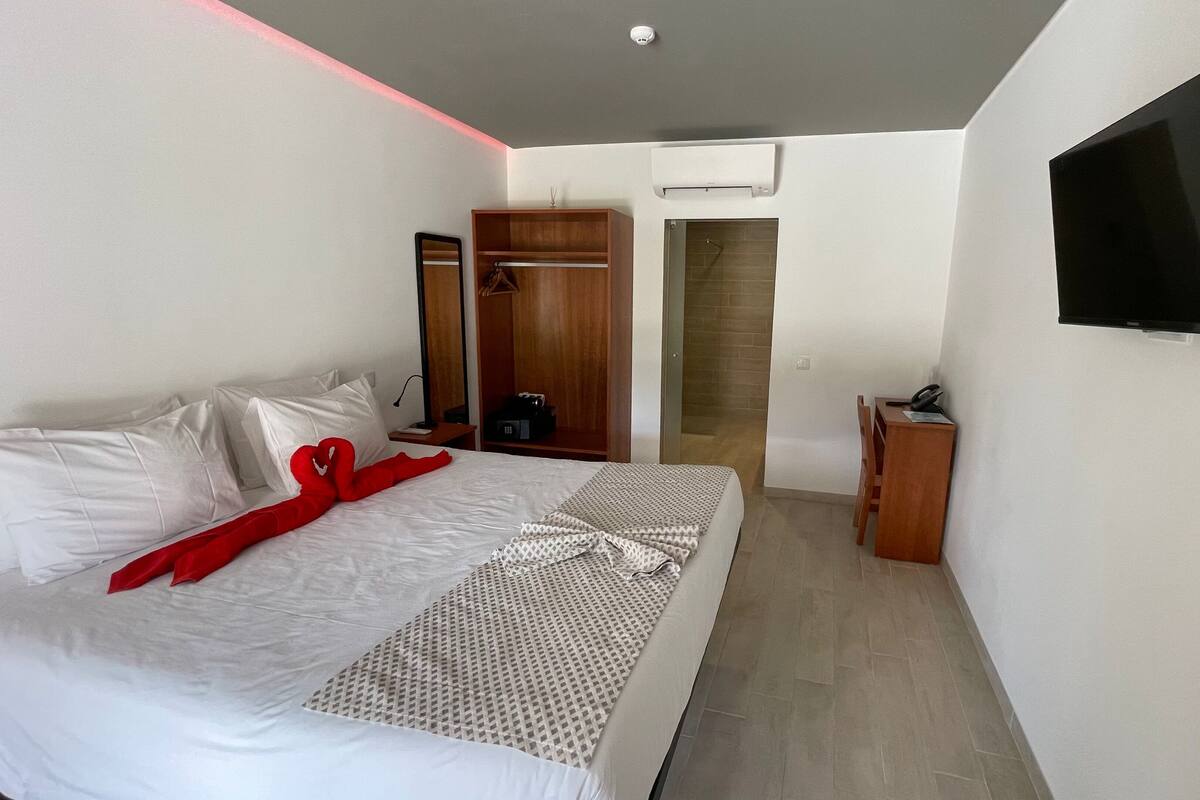 A modern hotel room with a neatly made bed, a TV mounted on the wall, a wooden wardrobe, and a small desk with a telephone.