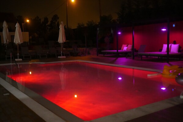 Zona de piscina nocturna con iluminación roja, sillones de descanso y sombrillas.