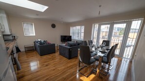 A modern living room with a sofa, dining table, and chairs. There is a TV and a clock on the wall. - The Coach House by Riis Property (Staines)