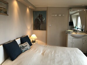 Et hotelværelse med seng, et natbord med lampe, et billede på væggen, et spejl over håndvasken og et vindue med gardiner.