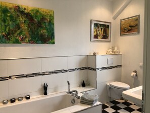 Et moderne badeværelse med badekar, toilet og h åndvask. Der hænger et stort abstrakt maleri over badekarret, og et mindre maleri over toilettet.