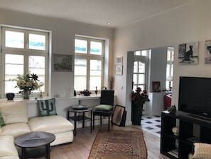 A living room with a sofa, a coffee table, a TV, and a fireplace. There are windows with curtains and a view of the outside.