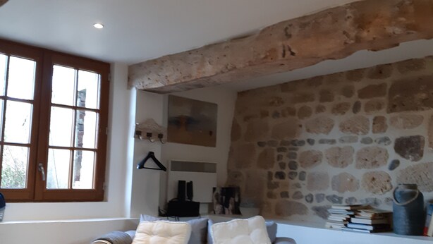 Ein gemütliches Wohnzimmer mit Steinwand, grauem Sofa mit Kissen, Holzbalkendecke und einem Fenster mit Jalousien.