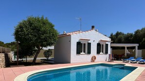 A swimming pool with a white building in the background, a tree, and a covered area with lounge chairs. - Villa with private pool in Cap d'Artrutx. (Cala en Bosc)