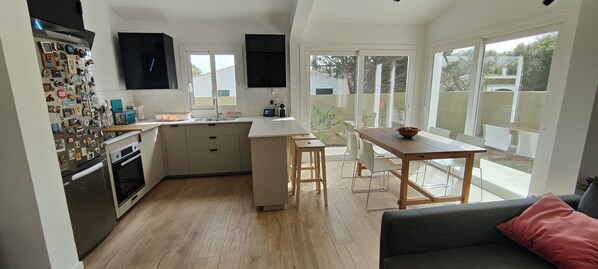 A modern kitchen with a central island, a dining area with a table and chairs, and large windows offering a view of the outdoors.