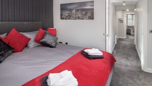 A neatly made bed with red and grey bedding, a cityscape painting on the wall, and a hallway leading to another room. - 2 Bedrooms Sleeps 6 Near Top Blackpool Attractions (Blackpool)