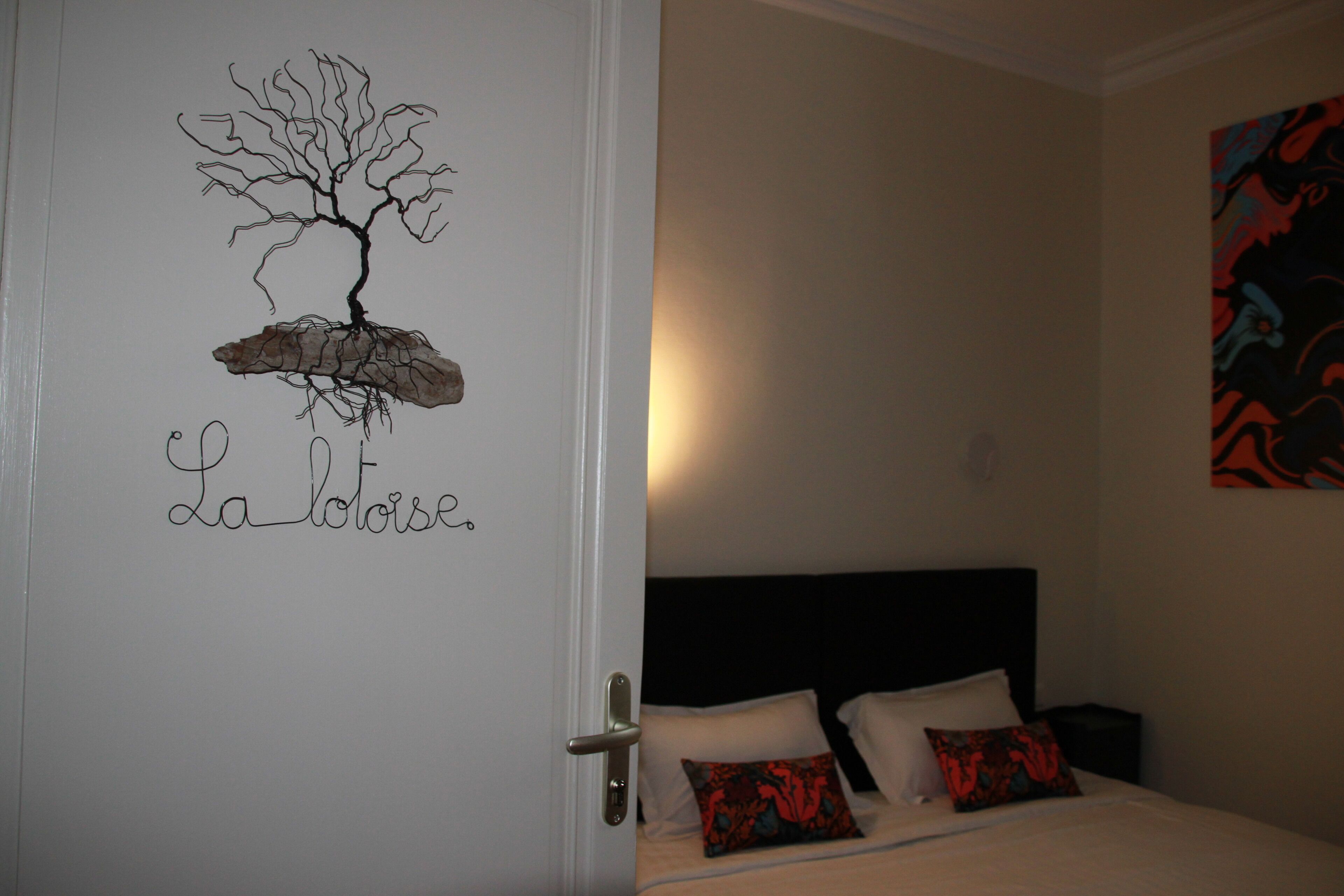 Habitación de hotel con cama, almohadas, puerta decorada con un árbol y texto, y una pared con un cuadro abstracto colorido.