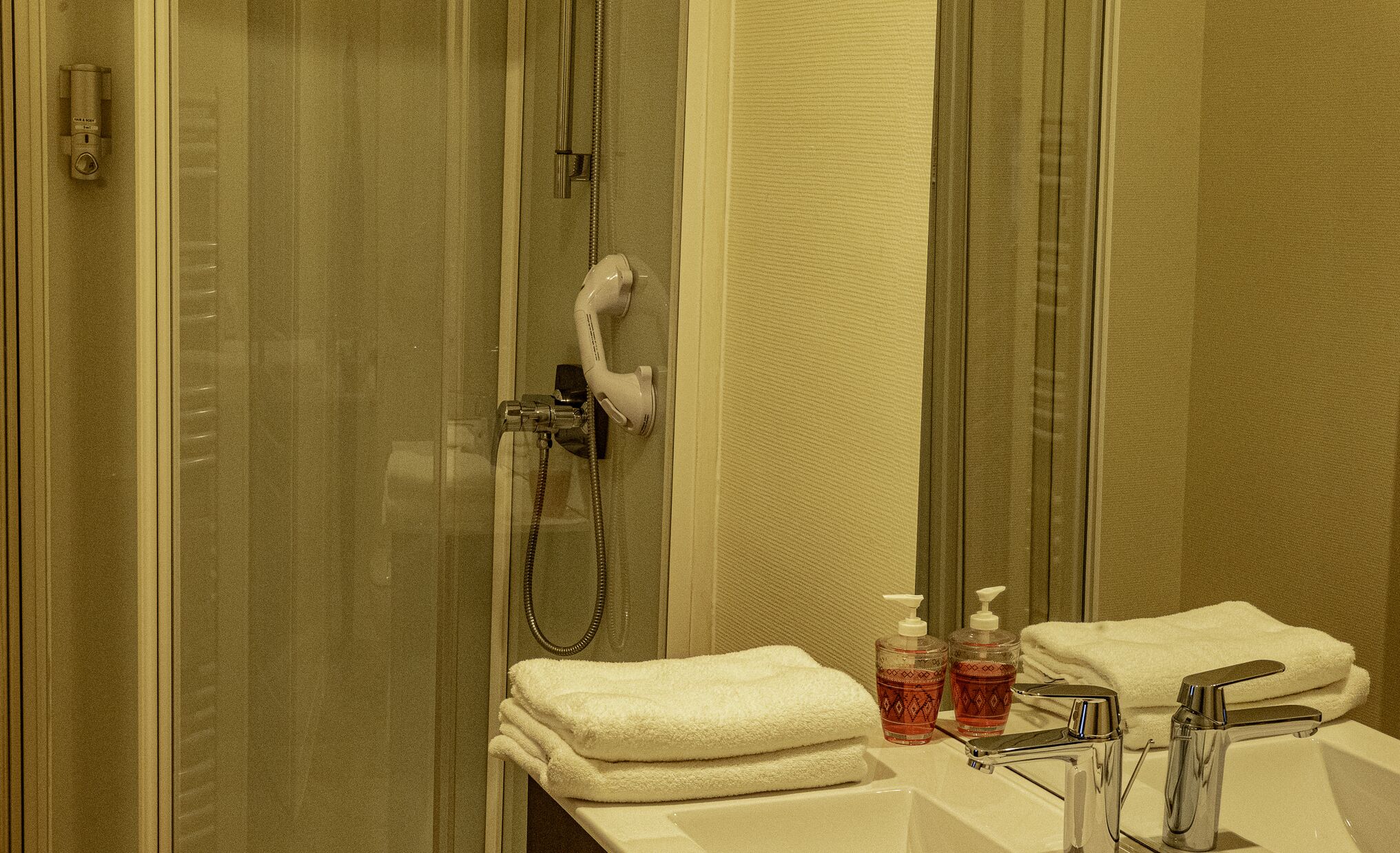 Un baño con cabina de ducha de vidrio, una toalla blanca, dos dispensadores de jabón y un lavabo con grifo cromado.