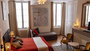 Habitación de hotel con una cama grande, un sofá, una mesita y una silla. Se observan ventanas con cortinas, un radiador y un espejo en la pared.