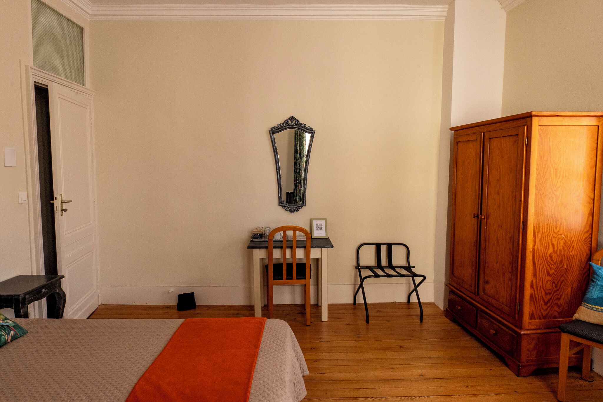 A bedroom with a bed, a wooden wardrobe, a desk with a chair, and a mirror on the wall.