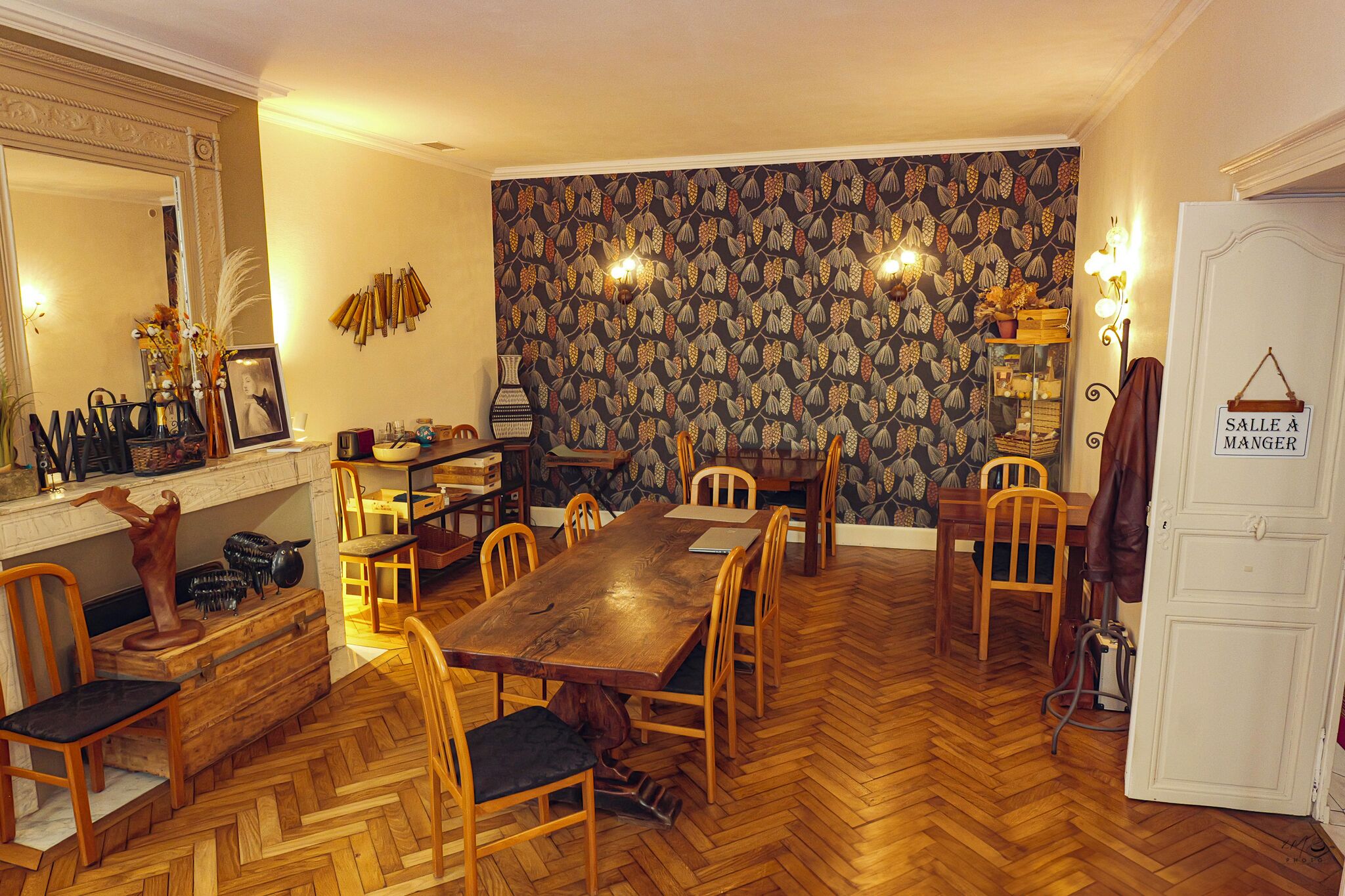 A dining room with a large wooden table and chairs, a patterned wallpaper, and a mirror on the wall.
