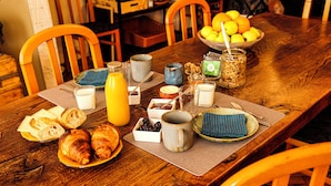 Una mesa de madera para el desayuno con croissants, pan, un bol de fruta y diversas bebidas.