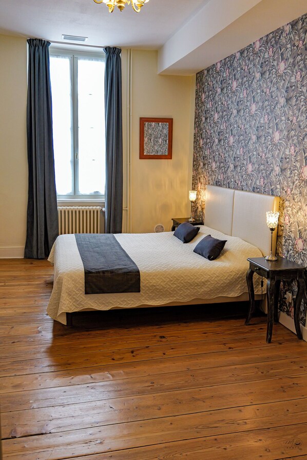 A bedroom with a large bed, wooden flooring, a radiator, a window with curtains, and floral wallpaper. - La Maison Duffour (Tonneins)