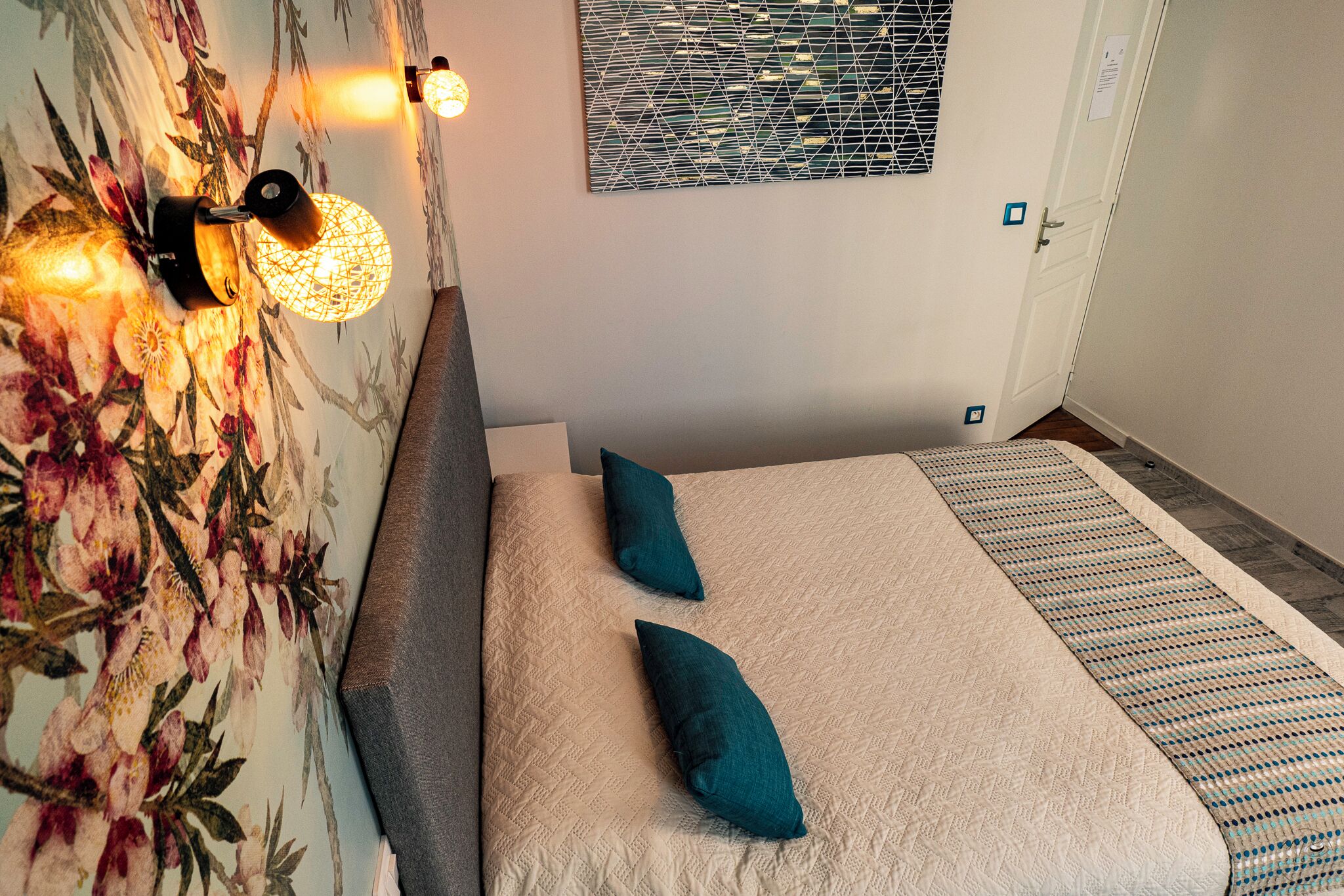 A bedroom with a floral wallpaper, a bed with a beige quilt and two teal pillows, a bedside table, and a wall-mounted light fixture.