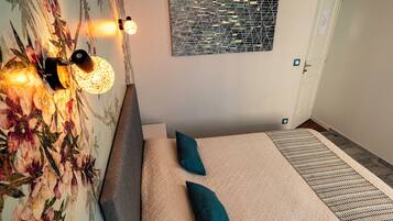 A bedroom with a floral wallpaper, a bed with a beige quilt and two teal pillows, a bedside table, and a wall-mounted light fixture.