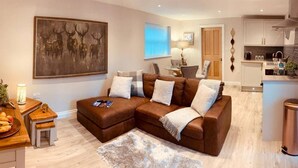 A modern living room with a brown sectional sofa, a dining table with chairs, a kitchen area, and a large painting on the wall. - Homely luxury cottage 2 (Tantobie)