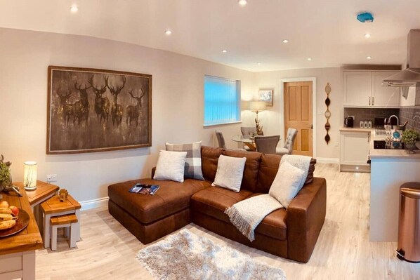 A modern living room with a brown sectional sofa, a dining table with chairs, a kitchen area, and a large painting on the wall.