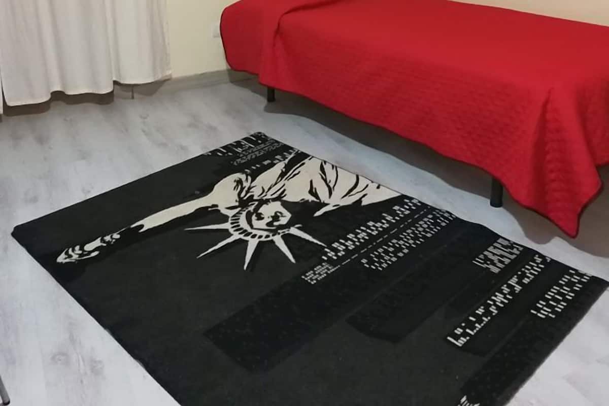 A room with a single bed covered in a red blanket, a black and white patterned rug, and a framed picture on the wall.