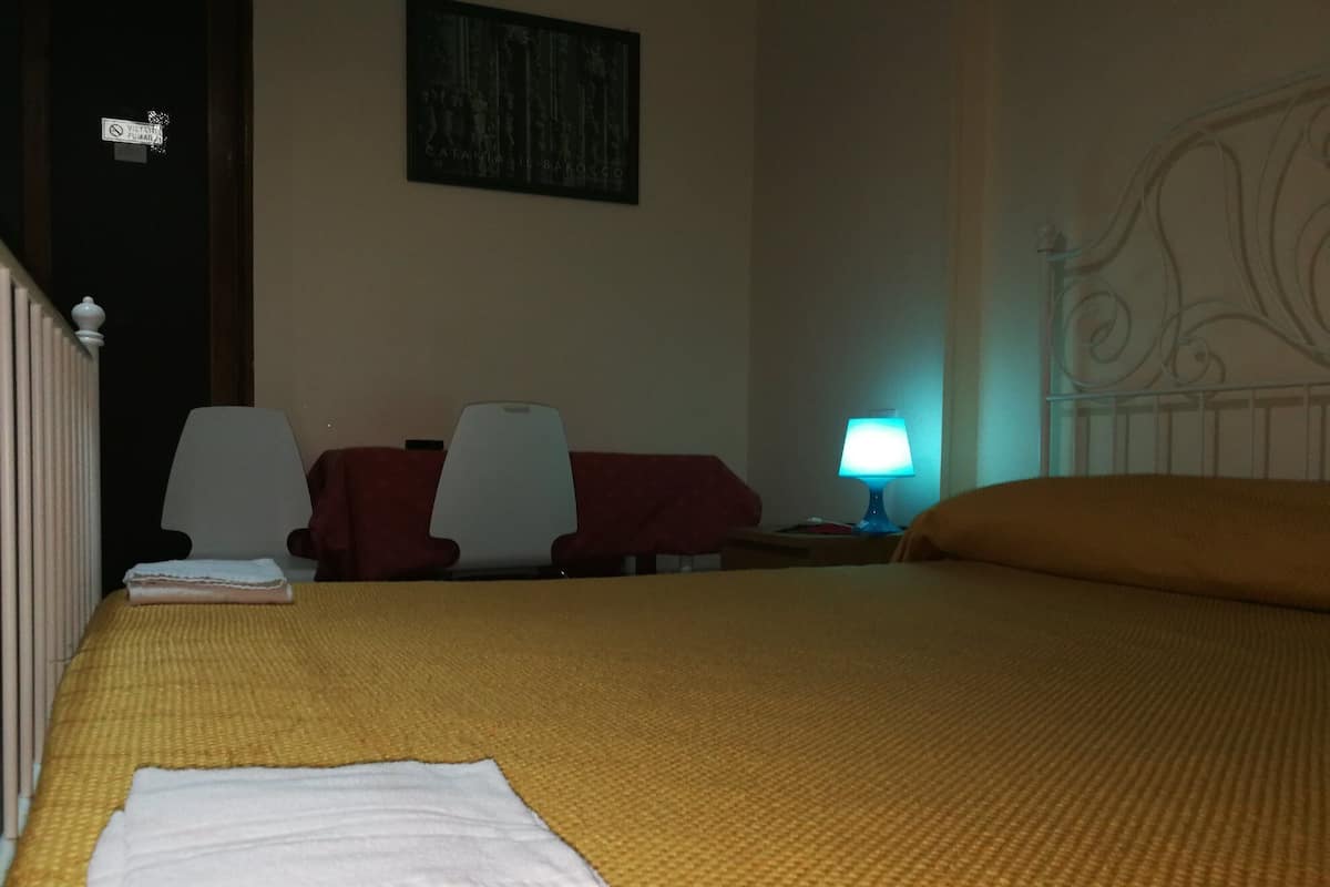 A hotel room with a bed, two chairs, a table, a nightstand with a lamp, and a door with a sign.