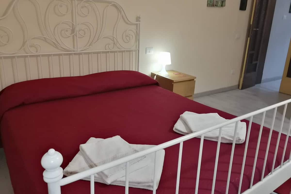 A neatly made bed with a red bedspread and white pillows, a bedside table with a lamp, and a door leading to another room.