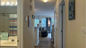 Um corredor com um banheiro à esquerda, uma sala de estar com um sofá e um abajur no centro, e uma porta com uma placa à direita.