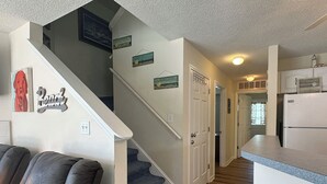 Interior de casa com escada que dá para um segundo andar, cozinha com geladeira e área de estar com sofá e um banquinho.