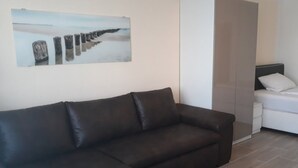 Un salón moderno con un sofá marrón oscuro, un armario blanco y una cama con ropa de cama blanca.