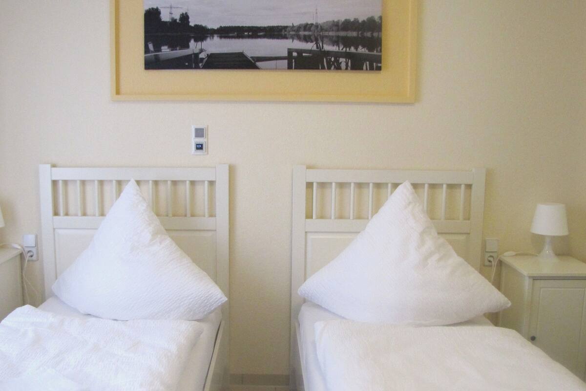 A hotel room with two single beds, white headboards, bedside lamps, a nightstand with a phone, and a framed picture on the wall.