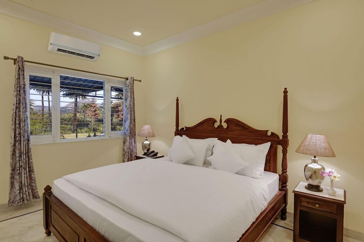 Quarto com cama de madeira com dossel, janela com cortinas e mesa de cabeceira com abajur.