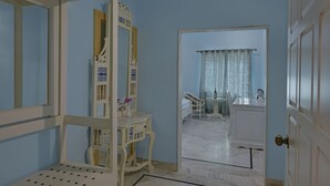 Habitación con tocador blanco, espejo y una silla. Se ve una puerta que da a otra habitación con un lavamanos y una silla.