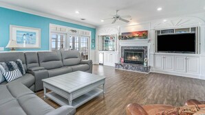 A living room with a grey sectional sofa, a white coffee table, a fireplace with a TV, and a ceiling fan. - All Aboard - Oceanfront in Garden City Beach (Garden City)