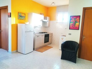 A compact kitchen with a refrigerator, stove, and cabinets, a small refrigerator, a chair, and a door.