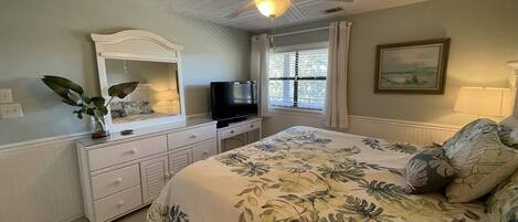 A bedroom with a bed, a ceiling fan, a dresser, a television, and a window with curtains.