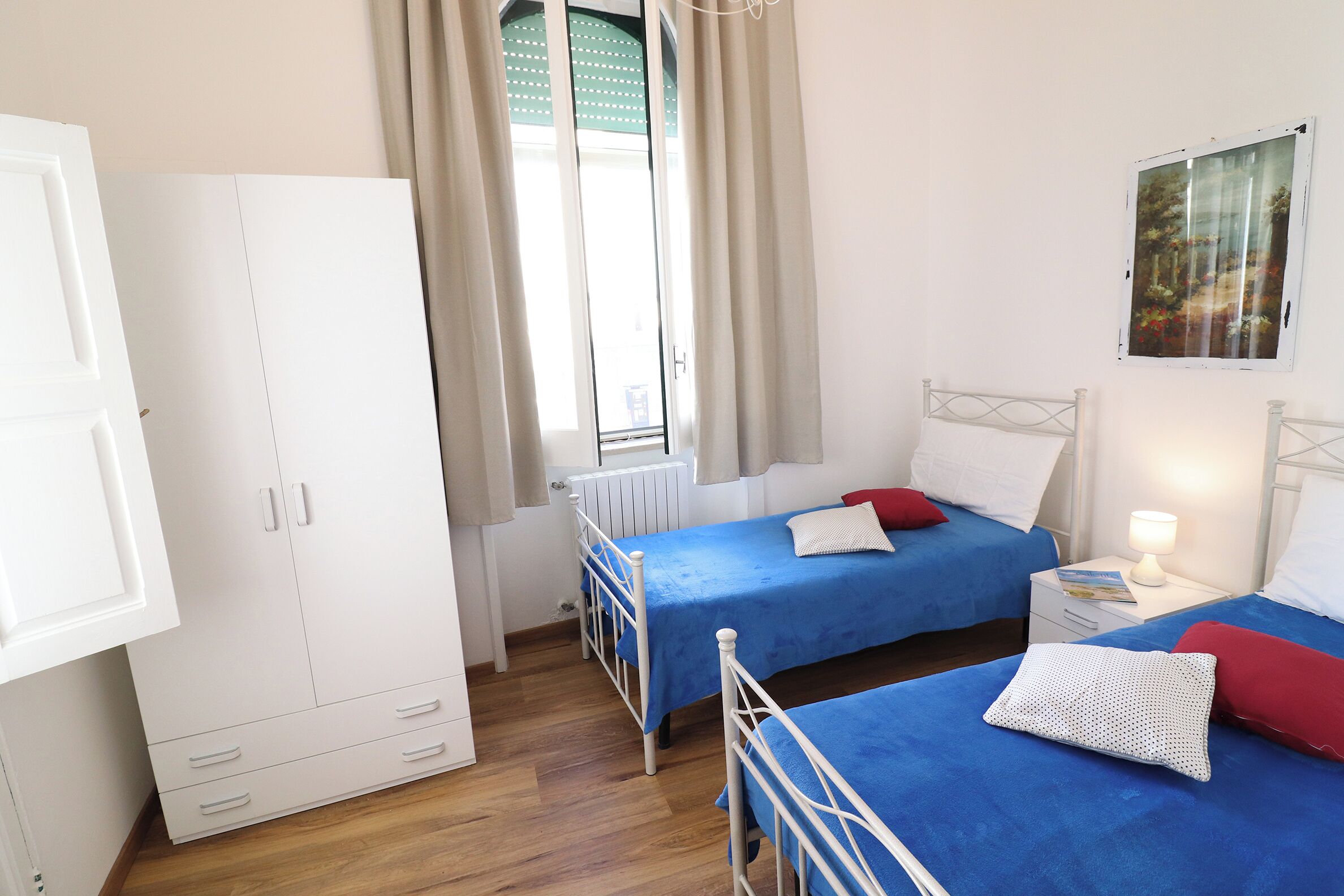 Une chambre avec deux lits, une armoire, une fenêtre avec des rideaux et un tableau au mur.