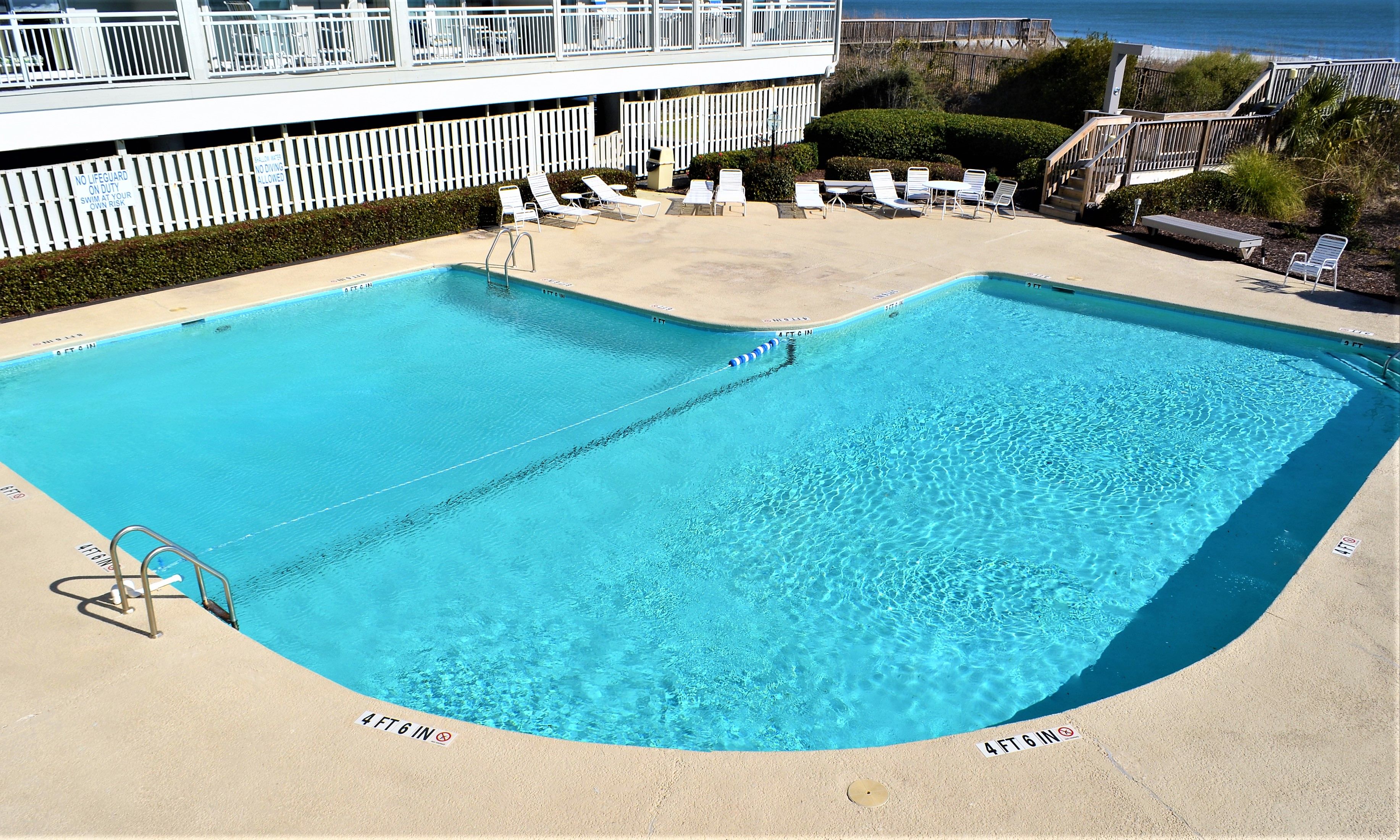 A rectangular pool with a diving board, surrounded by lounge chairs and a white fence.