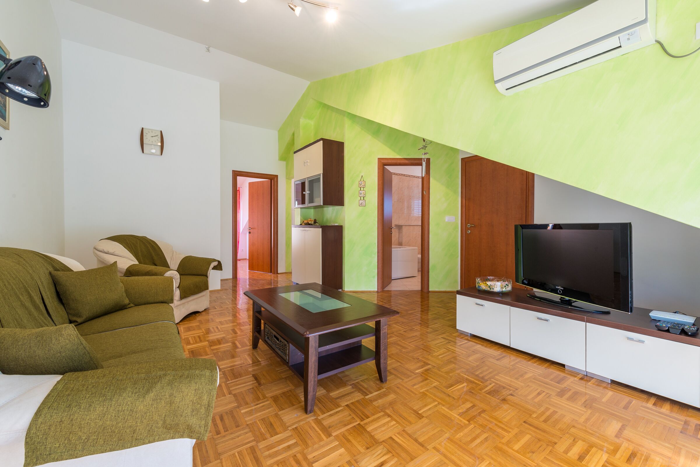 Una sala de estar con una pared verde de acento, piso de madera, un sofá, una mesa de centro, un televisor y un área de cocina con armarios y un microondas.