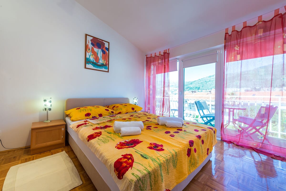 A bedroom with a bed, a nightstand, a window with curtains, and a view of the outside.