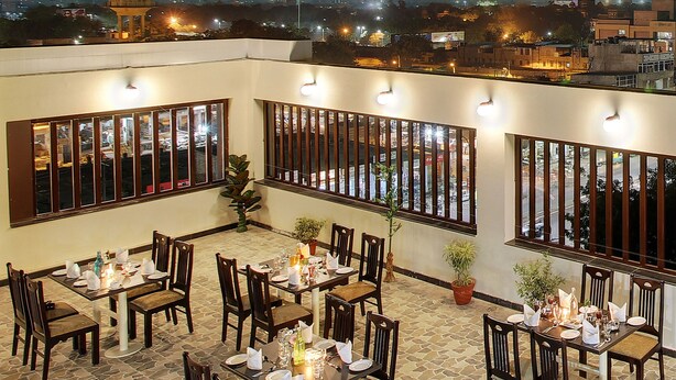 An outdoor dining area with multiple tables and chairs, large windows with blinds, and a view of the cityscape at night. - Trulyy Rudransh inn (Jodhpur)