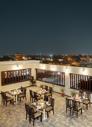 Un área de comedor al aire libre con varias mesas y sillas, ventanas grandes con persianas y vista a la ciudad iluminada de noche.