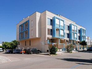 Ein modernes Gebäude mit beiger Fassade und blauen Akzenten, mit mehreren Balkonen und einem Parkplatz mit einigen Autos.