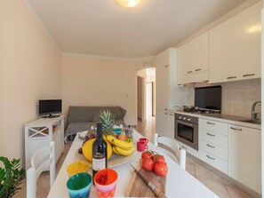 Eine moderne Küche mit weißen Schränken, einem Herd und einem Fernseher. Es gibt einen Esstisch mit einer Weinflasche, Obst und bunten Tassen.