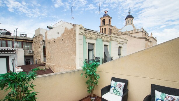 Un balcón con dos sillones de mimbre, una planta en maceta y un edificio con una torre de campanas al fondo.