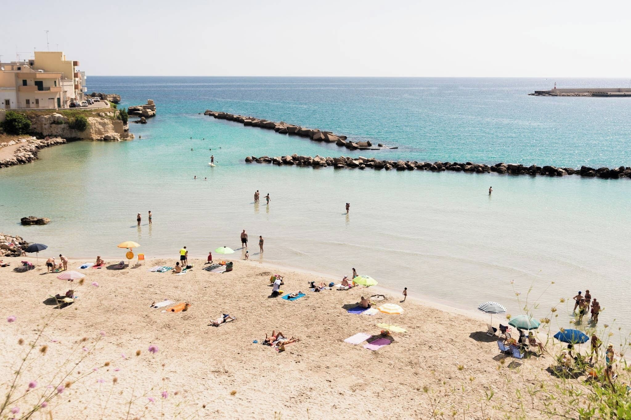 Une plage où l’on voit des gens prendre des bains de soleil et se baigner, une digue rocheuse et un bâtiment situé sur la gauche.