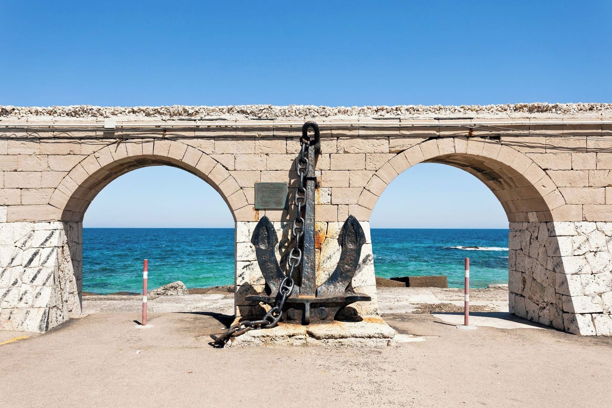Un arco de piedra con una escultura de ancla en el centro, con vistas a la costa.